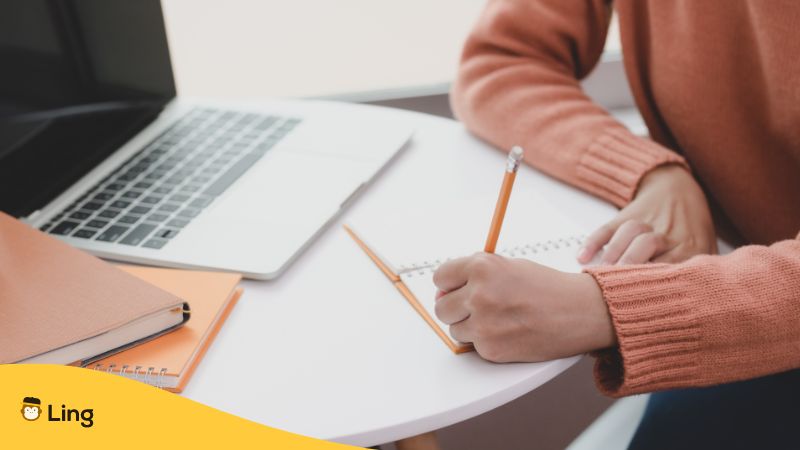Frau sitzt am Tisch und schreibt in ein Notizbuch, vor sich ist ein aufgeklappter Laptop