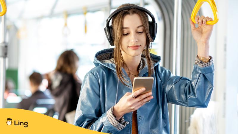 Junge Frau steht in der Bahn und hält sich mit einer Hand fest und in der anderen hat sie ihr Handy und lernt beim Pendeln mit der Sprachlern-App Ling eine neue Sprache